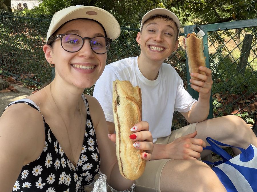 Hugo et Camille souriants, un sandwich à la main, assis dans un parc.